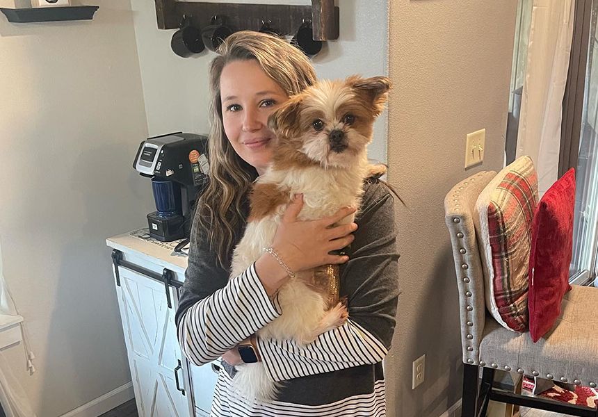 A person smiling while holding a small, fluffy dog indoors near a coffee maker.