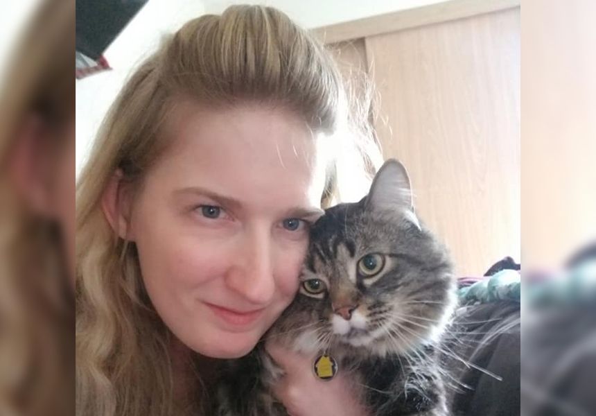 A person with long blond hair smiles while holding a fluffy, striped cat close to their face.