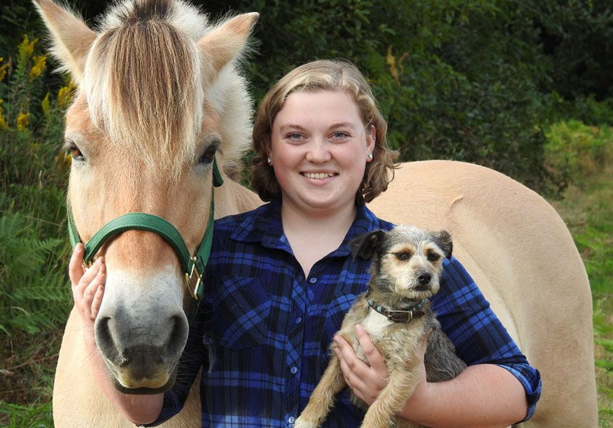 Person smiling while holding a small dog and standing beside a horse.