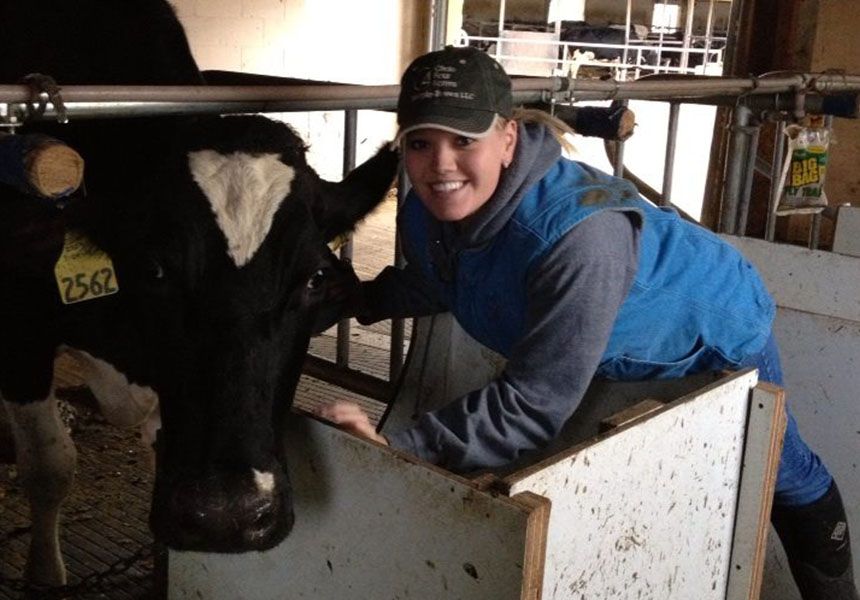 A person wearing a cap is smiling and standing beside a cow in a pen.