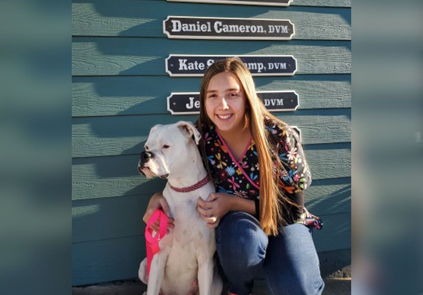A woman smiling next to a white dog with a pink collar, crouched in front of a wall with veterinary name plaques.