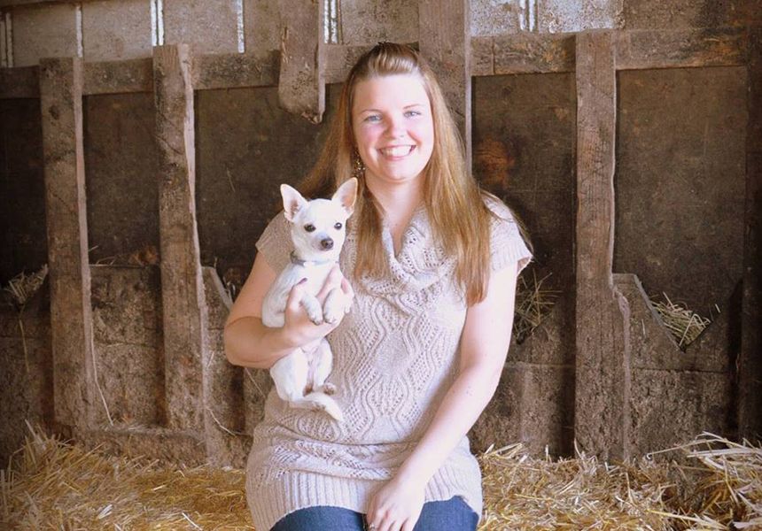 A woman sitting and smiling, holding a small dog, in a rustic setting.