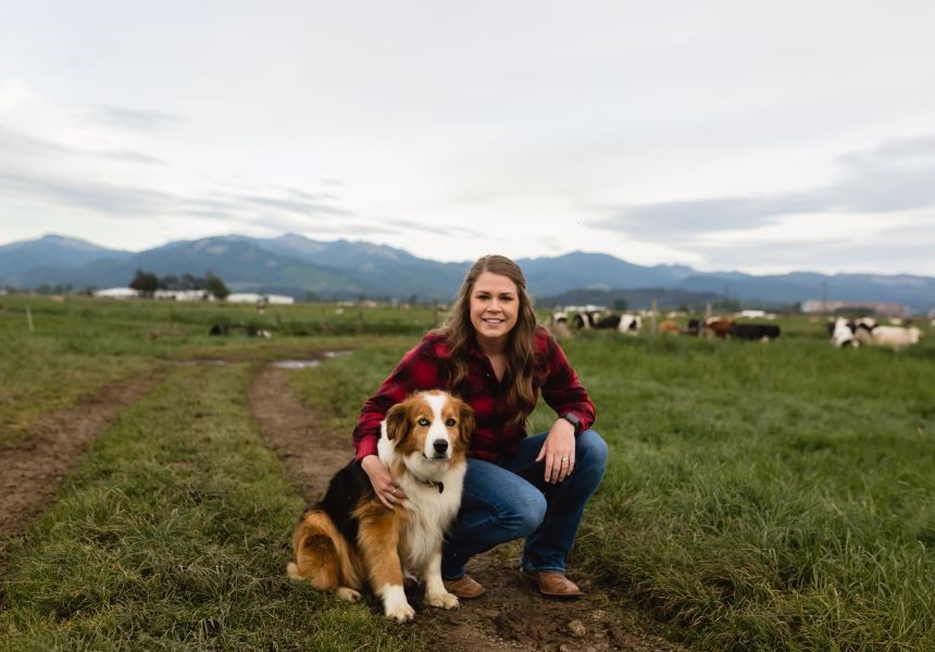 Woman kneeling beside a dog on a grassy field with cows in the background under a cloudy sky.