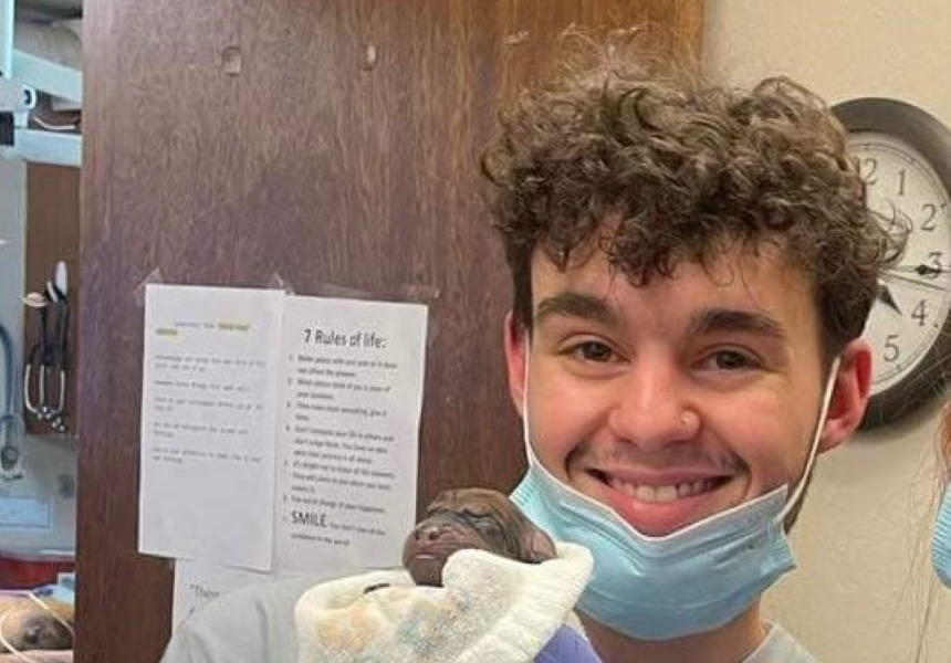 A person with curly hair and a face mask smiles while holding a small, newborn animal wrapped in a towel.